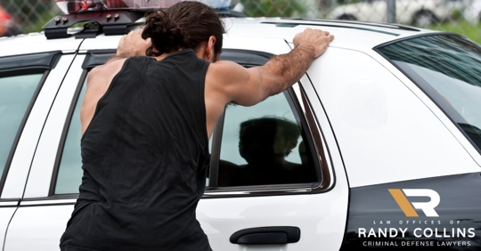 man in tank top with hands up on police car facing away from the camera